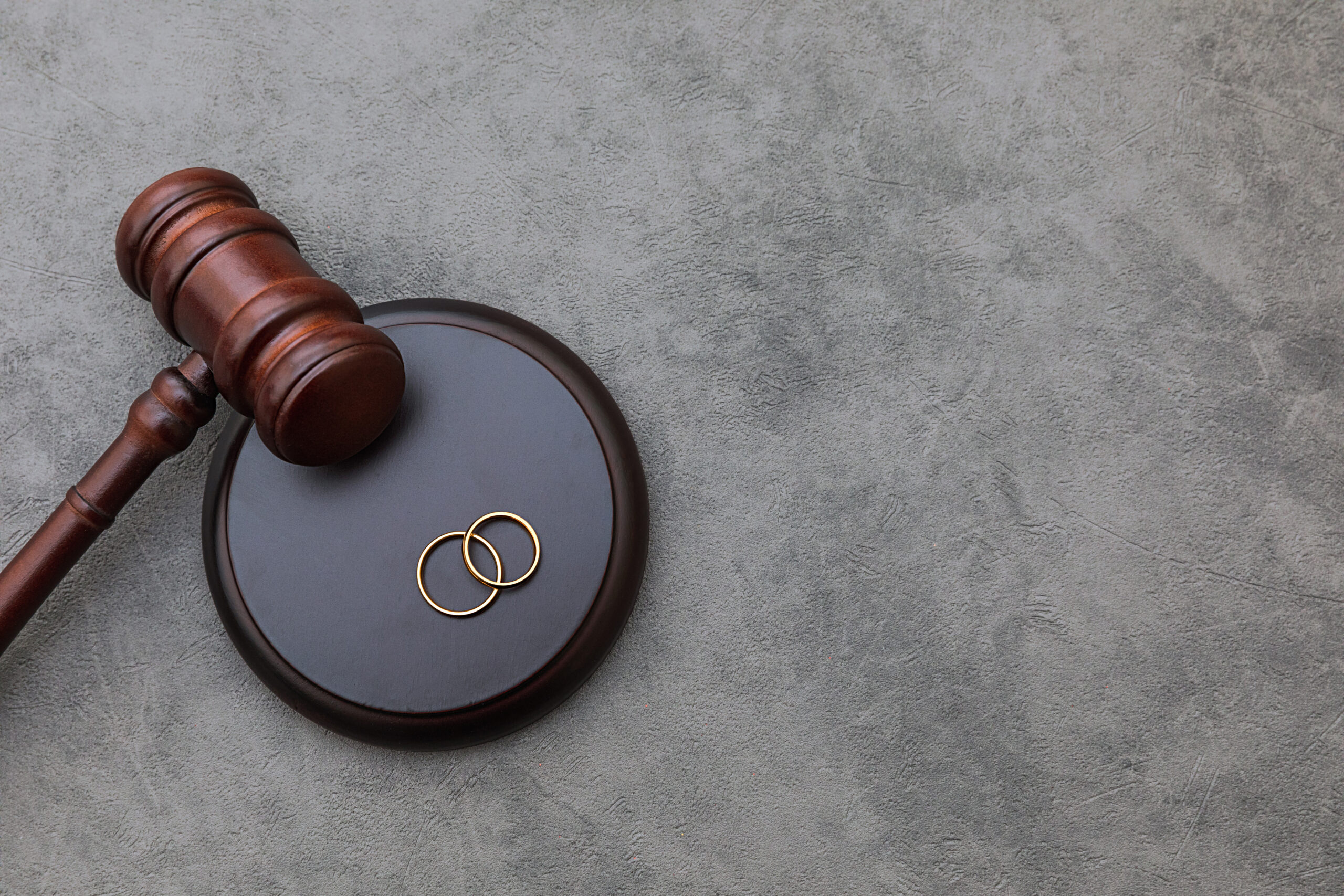 A wooden gavel rests on a round block next to two intertwined wedding rings on a gray surface, symbolizing legal aspects of marriage.