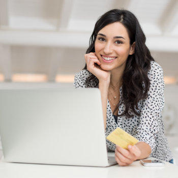Woman smiling at the camera while holding a credit card and sitting in front of a laptop.