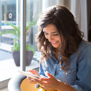 Woman in denim shirt smiling while using a tablet by a window.