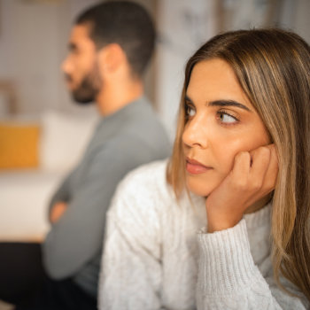A woman looking thoughtful with a man sitting in the background out of focus.