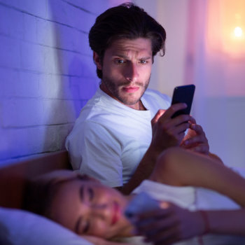 Man using smartphone in bed next to sleeping woman at night.