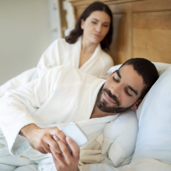 A man wearing a sleep mask and a bathrobe checks his phone while lying in bed, with a woman sitting up behind him.