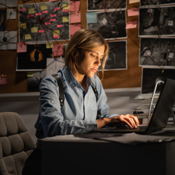 Woman working intently on a laptop at a desk with a bulletin board covered in notes and photos behind her.