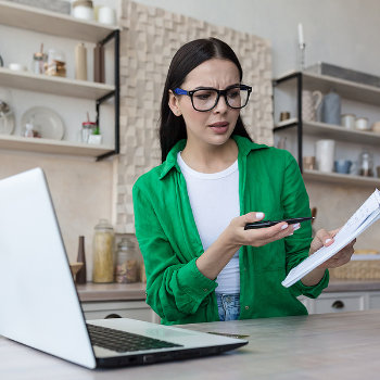 Woman in green jacket working from home with laptop, looking perplexed at a document and holding a smartphone.